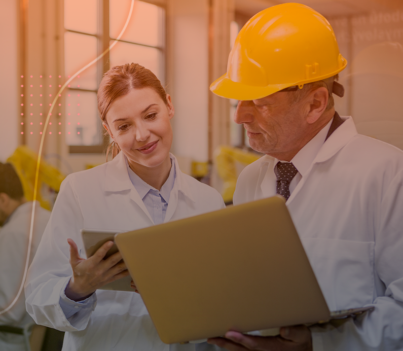 A female engineer and a male supervisor, both in safety gear, collaborate on a laptop and tablet in a manufacturing environment, discussing project details with smiles.